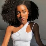 Femme habillé en blanc et assise sur un lit dans une chambre au mur blanc portant une perruque afro aux cheveux naturels et crépus.