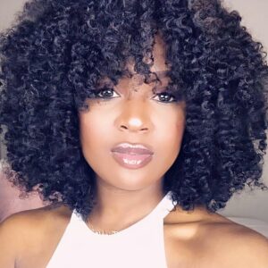 Femme avec un haut blanc pose dans une chambre assise sur un lit. Elle porte une perruque afro coupe naturelle de couleur noire.
