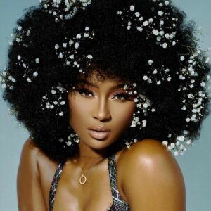 Femme en débardeur devant un mur bleu portant une perruque afro naturelle crépue et bouclée noire. La perruque est décorée avec des petites fleurs blanches.