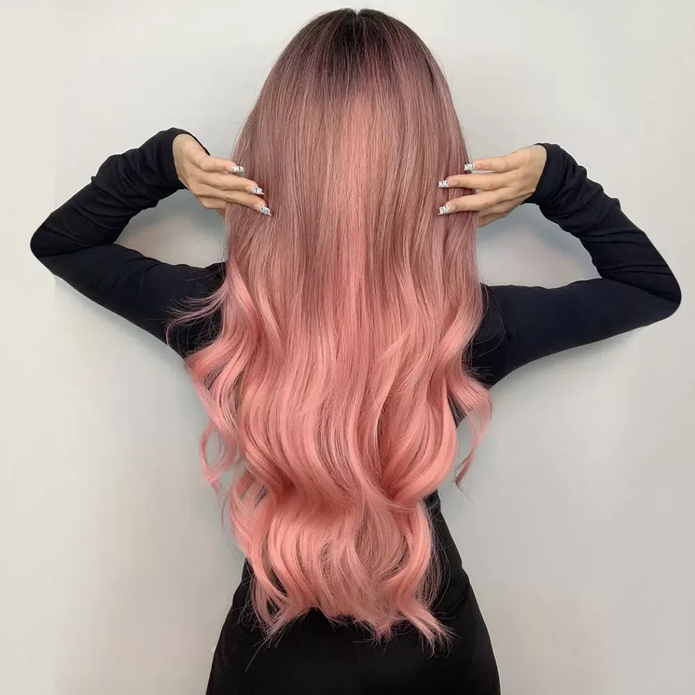 perruque longue rose ondulée portée par une femme de dos en robe moulante noir sur fond gris avec mains sur les cheveux