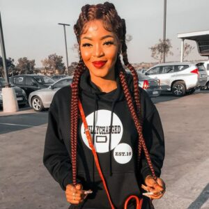 Femme sur un parking avec des voitures et un bâtiment en arrière plan. Elle porte un pull à capuche noir et blanc et tient les tresses de sa perruque tresses africaines en fibre synthétique rousse.